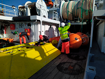 Fortøjningsarbejde på R/V Jákup Sverri i maj 2021. Alle strømmålere er hentet op og ligger på dækket. Der gøres klar til transport til Havstovan, hvor data læses og friske batterier sættes i til nye udlægninger. Forrest en strømmåler i trawlsikret ramme (gul), som har været udlagt på Island-Færø ryggen og bagved de andre strømmålere i orange flotation bøjer ( Karin M. H. Larsen).
